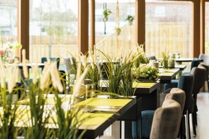 Un restaurant où les tables sont dressées pour le service, agrémenté de plantes en pot et offrant une vue sur l’extérieur.