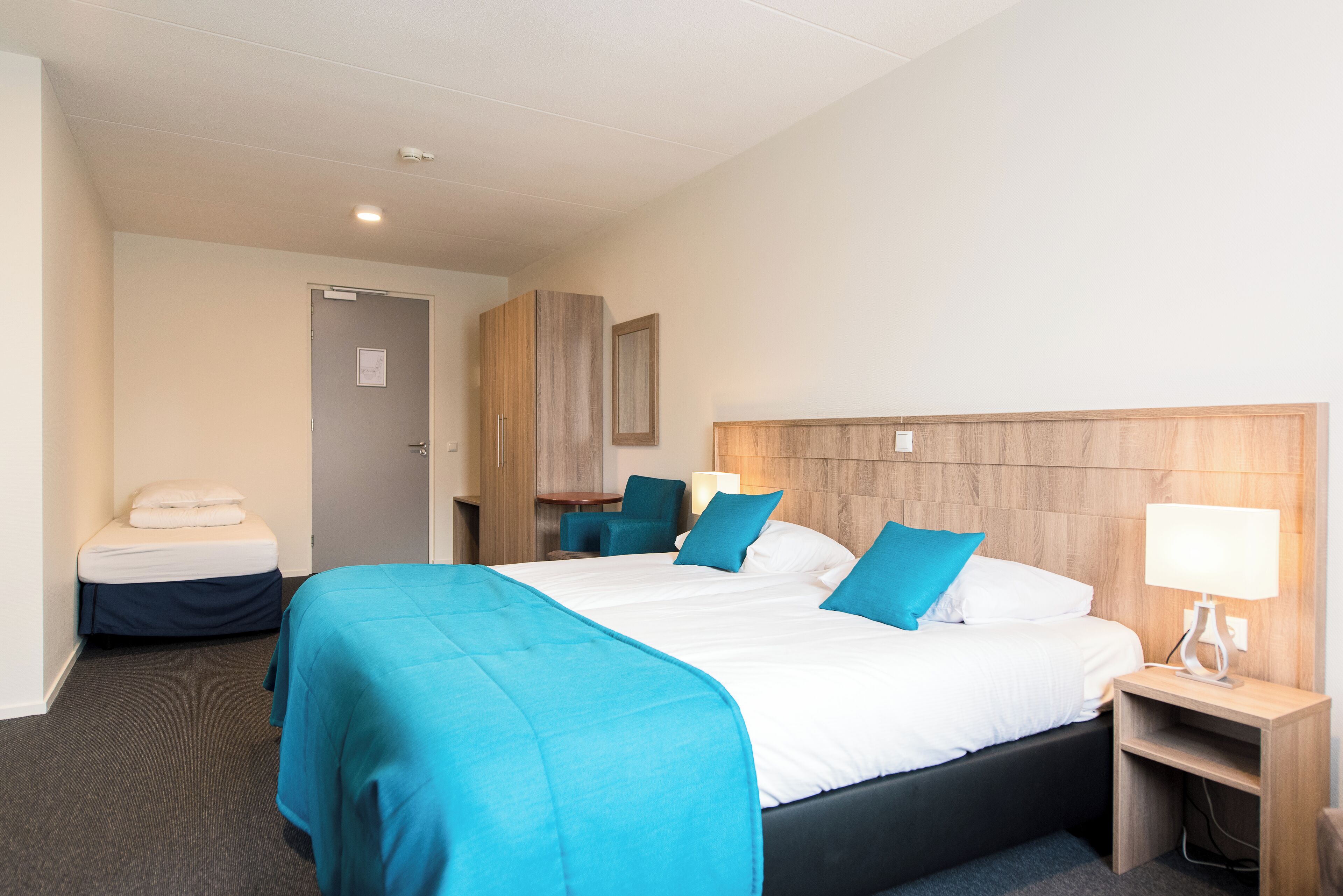 Habitación de hotel con una cama grande, una mesita de noche con lámpara, un escritorio con silla y un armario.