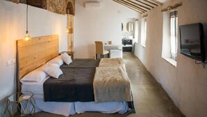 Un dormitorio con cabecera de madera, cama con cubierta oscura, mesita de noche pequeña y una silla. Se observa un escritorio y un espejo al fondo.
