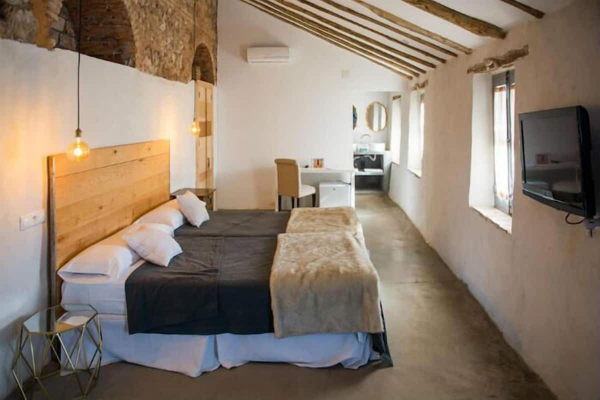 Un dormitorio con cabecera de madera, cama con cubierta oscura, mesita de noche pequeña y una silla. Se observa un escritorio y un espejo al fondo.