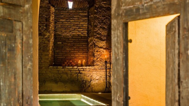 Habitación con paredes de piedra, una pequeña ventana, una escalera que sube a un nivel superior y una vela en el suelo.