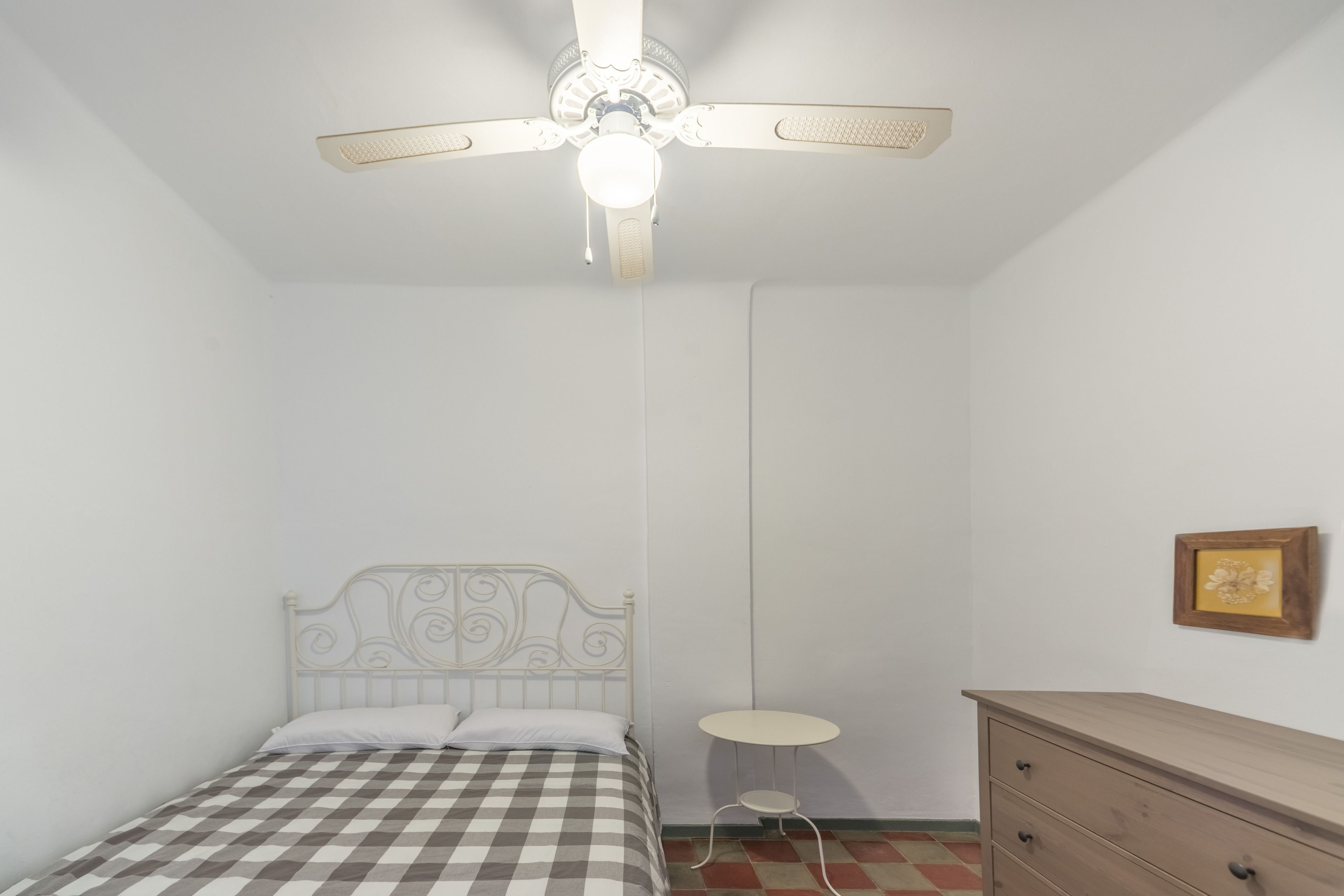 Een slaapkamer met een bed, een plafondventilator, een klein rond tafeltje en een dressoir.