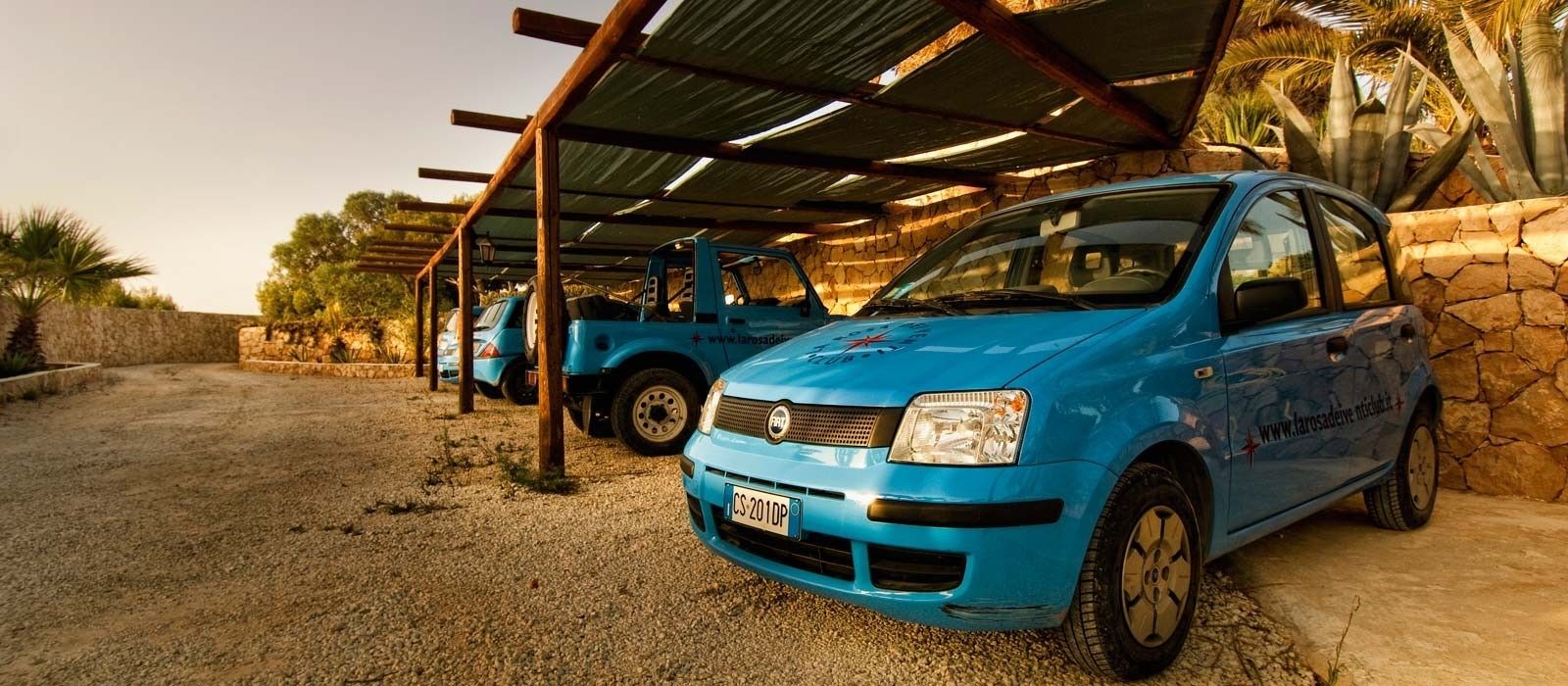 Ein blaues Auto steht unter einer Überdachung mit einer Steinmauer und Palmen im Hintergrund.