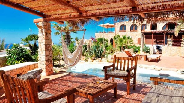 A poolside area with wooden furniture, a hanging hammock, and a stone building in the background. - La Rosa Dei Venti (Buggerru)