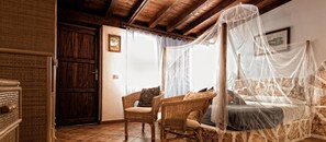 A rustic bedroom with a stone wall, a wooden door, a wicker chair, a bed with a canopy, and a framed picture on the wall. - La Rosa Dei Venti (Buggerru)