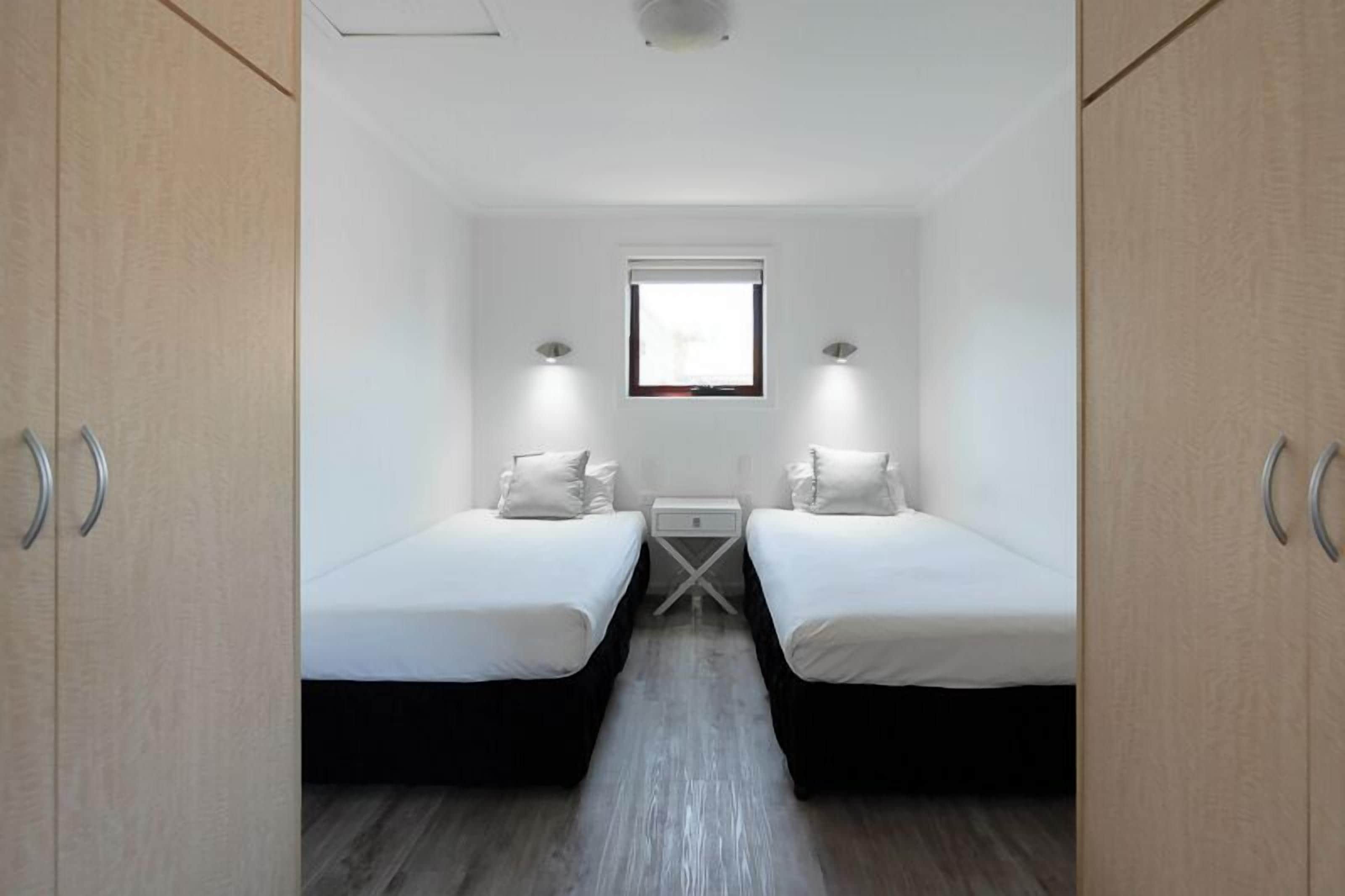 Une chambre de taille modeste, équipée de deux lits simples, d’une petite table de chevet, d’une fenêtre et d’un luminaire au plafond.