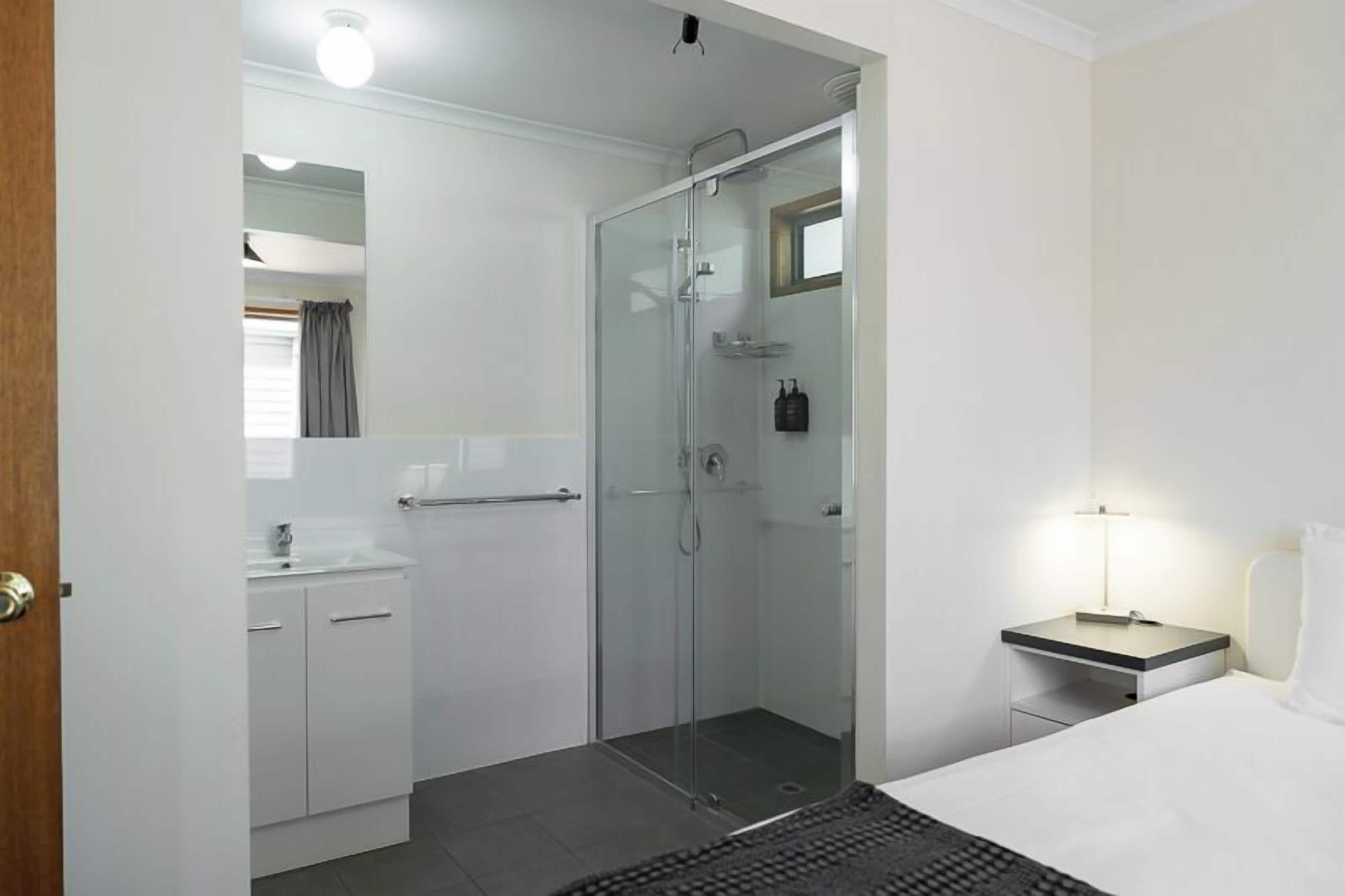 A modern bathroom with a glass shower enclosure, a white sink, and a bedside table with a lamp.