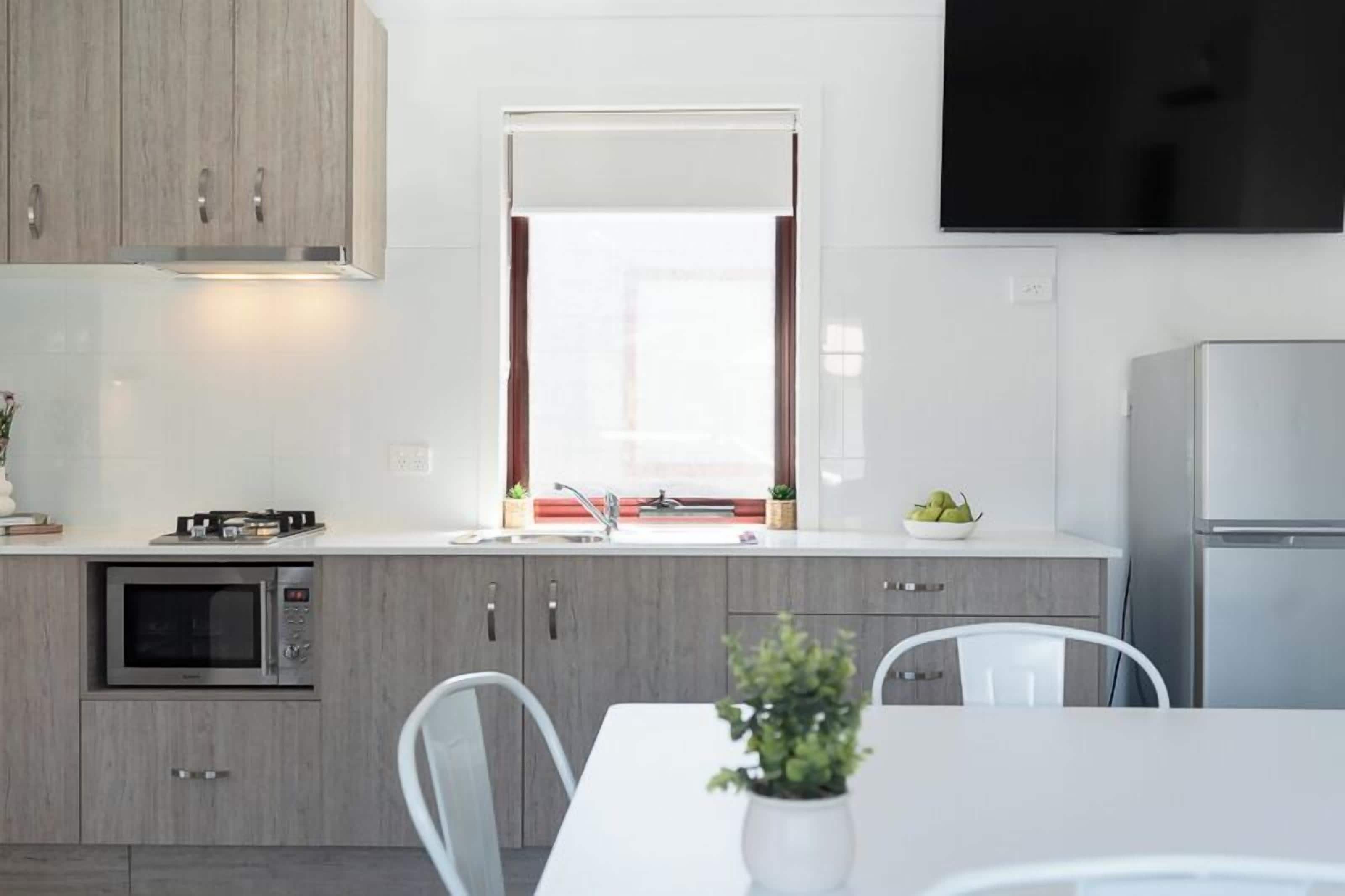 A modern kitchen with a built-in microwave, a gas stove, and a sink. There is a window with curtains, a flat-screen TV mounted above the counter, and a refrigerator to the right.