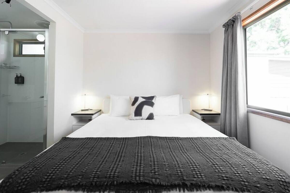 A hotel room with a large bed, two bedside tables, a nightstand, a window with curtains, and a bathroom with a glass shower enclosure.