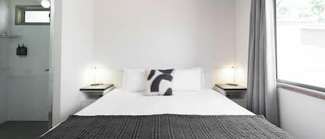 Una habitación de hotel con una cama grande, dos mesitas de noche, un tocador, una ventana con cortinas y un baño con cabina de ducha de vidrio.