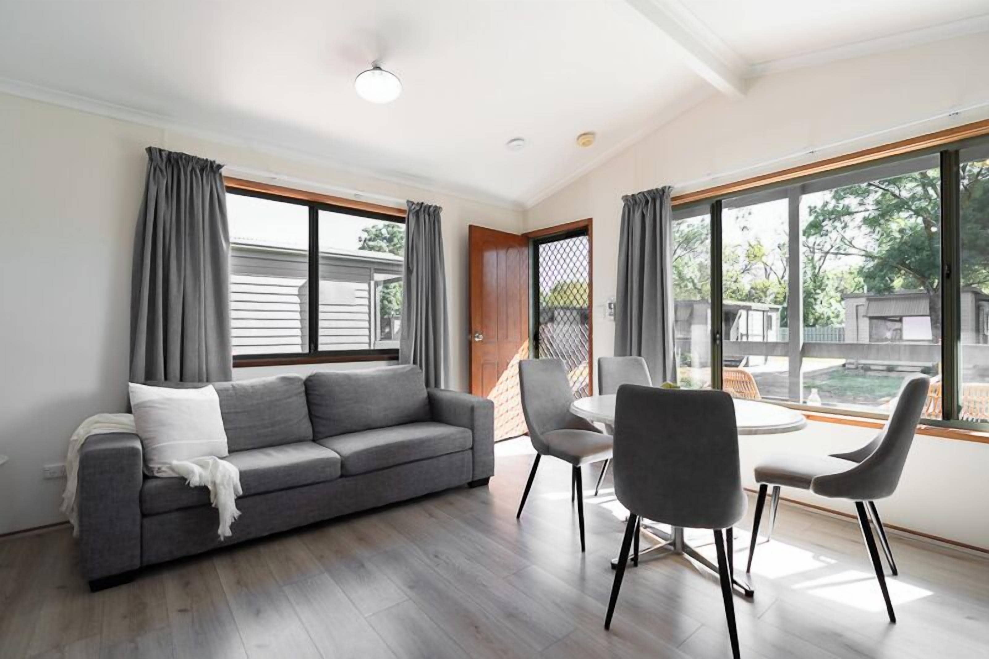 Een moderne woonkamer met een grijze bank, twee fauteuils, een klein rond tafeltje en grote ramen met grijze gordijnen.