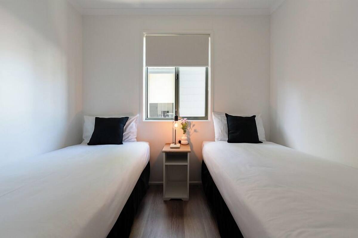 A small, clean room with two single beds, a nightstand with a lamp, and a window providing natural light.
