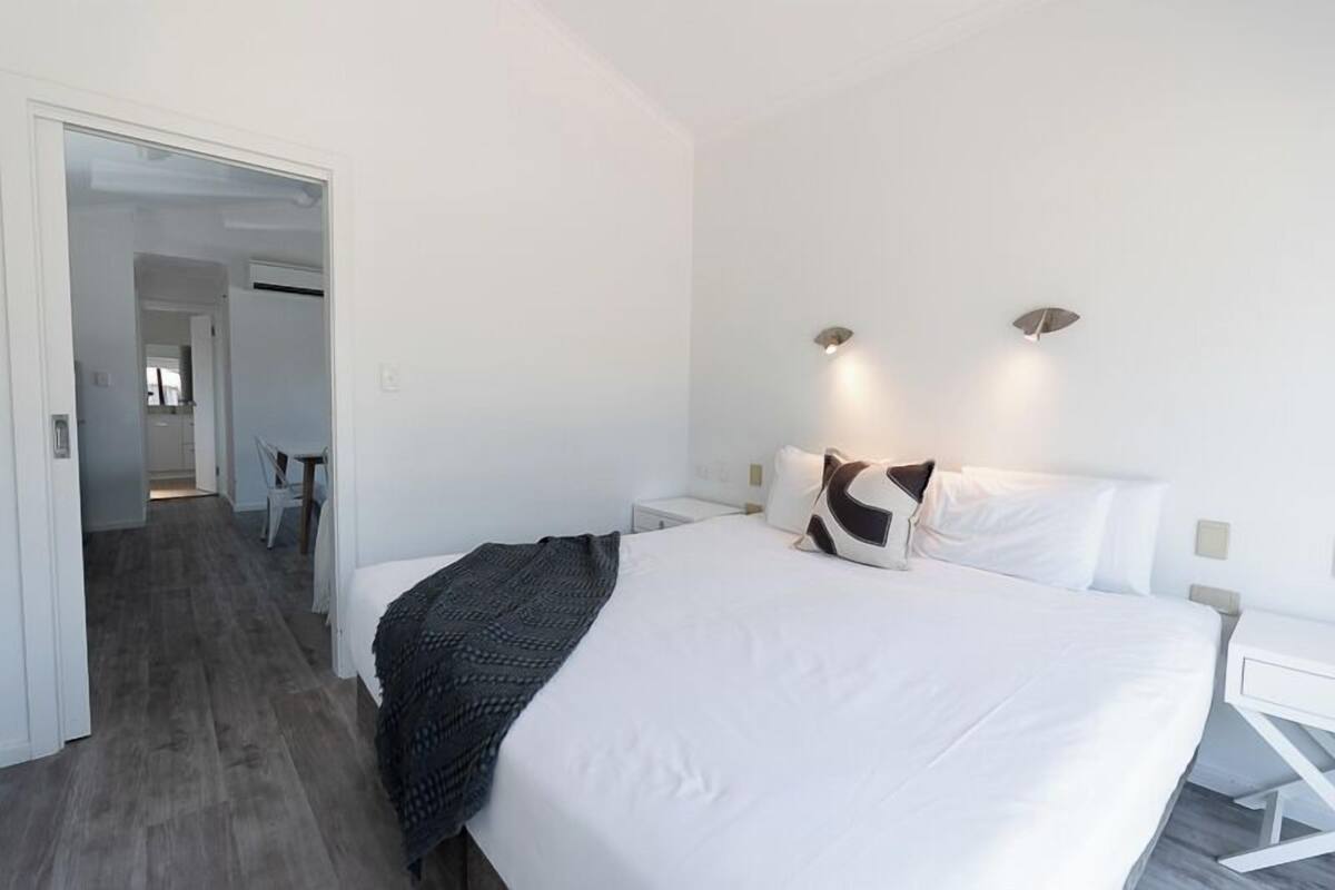 A modern hotel room with a large bed, white walls, and a wooden floor. There is a desk with a chair and a small table with a chair in the corner.