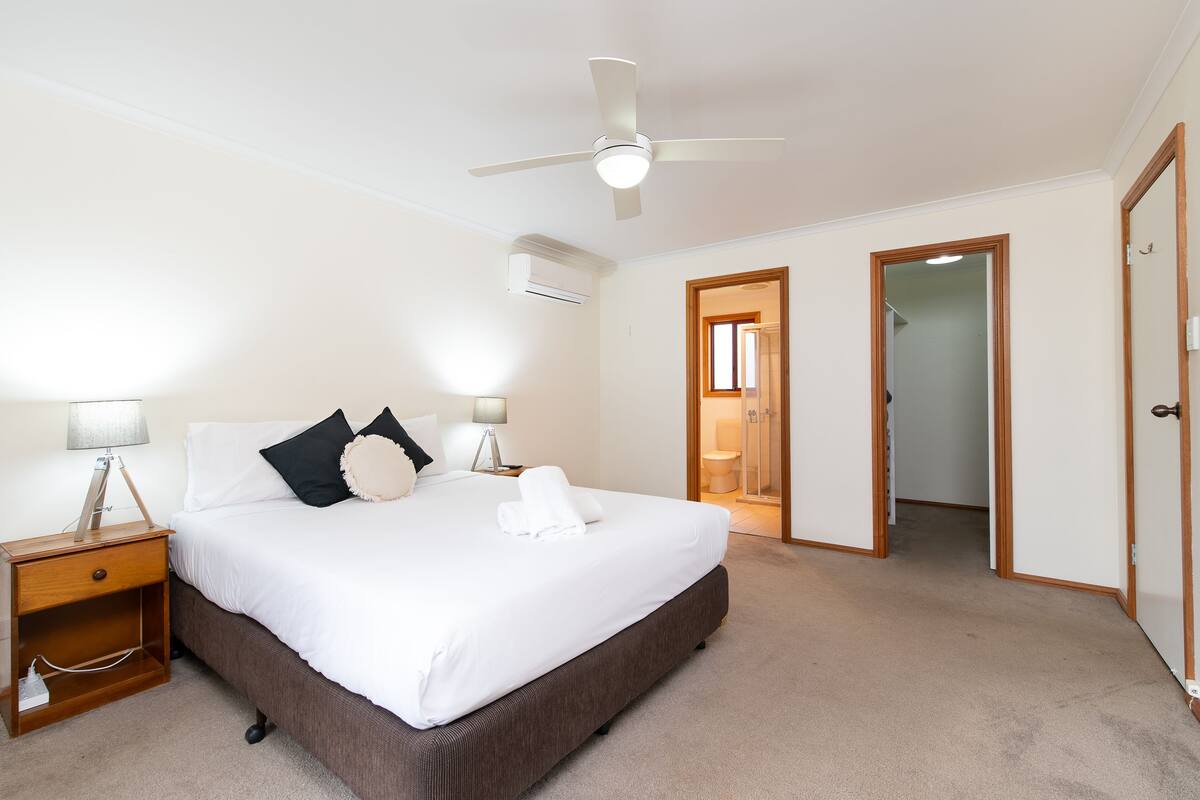 A bedroom with a bed, bedside tables, a ceiling fan, and an air conditioning unit.