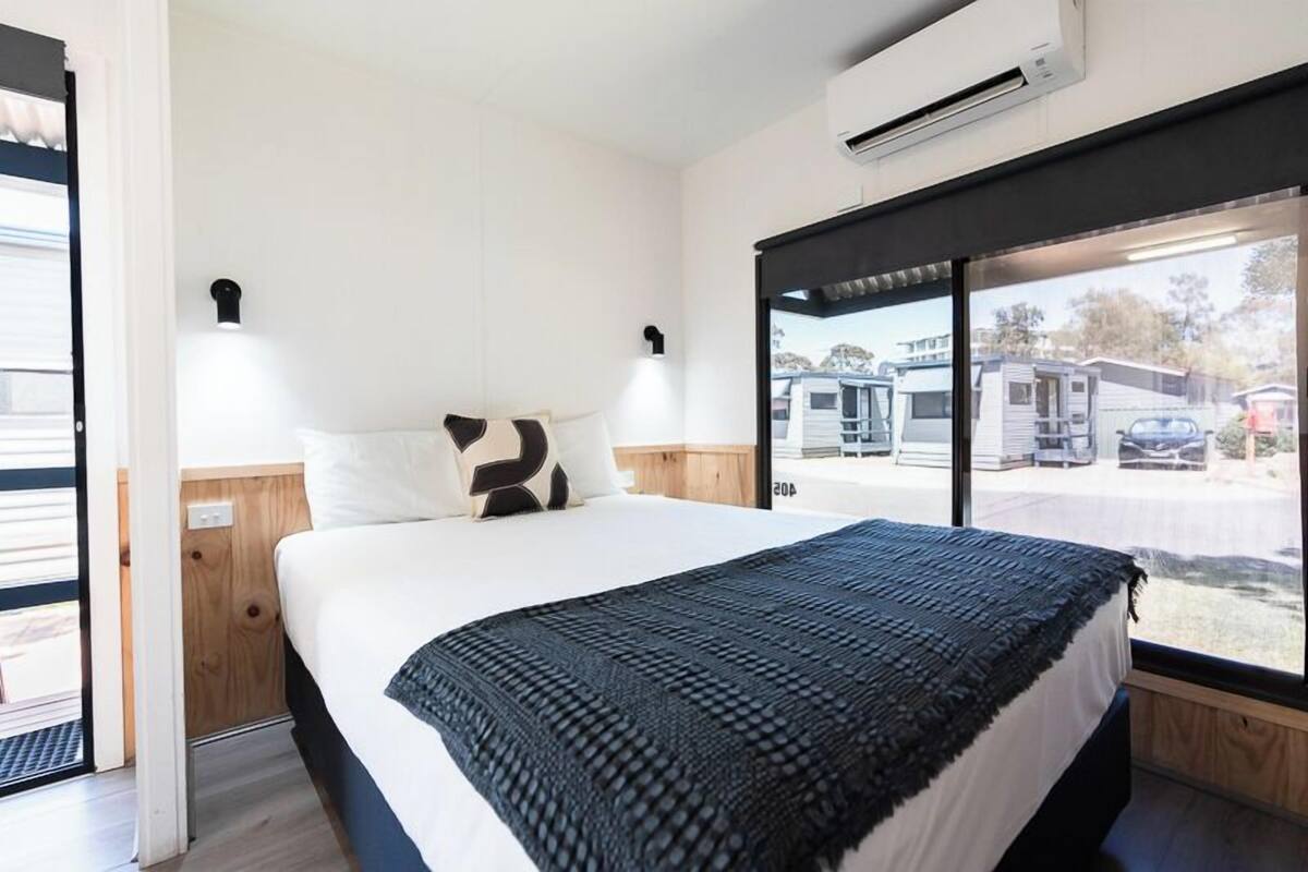 A bedroom with a bed, a window with a view of parked vehicles, and an air conditioning unit.