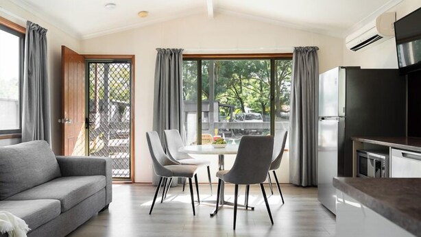 A modern living room with a sofa, dining table, chairs, and a kitchen area with a refrigerator and microwave. - Adelaide Caravan Park (Hackney)