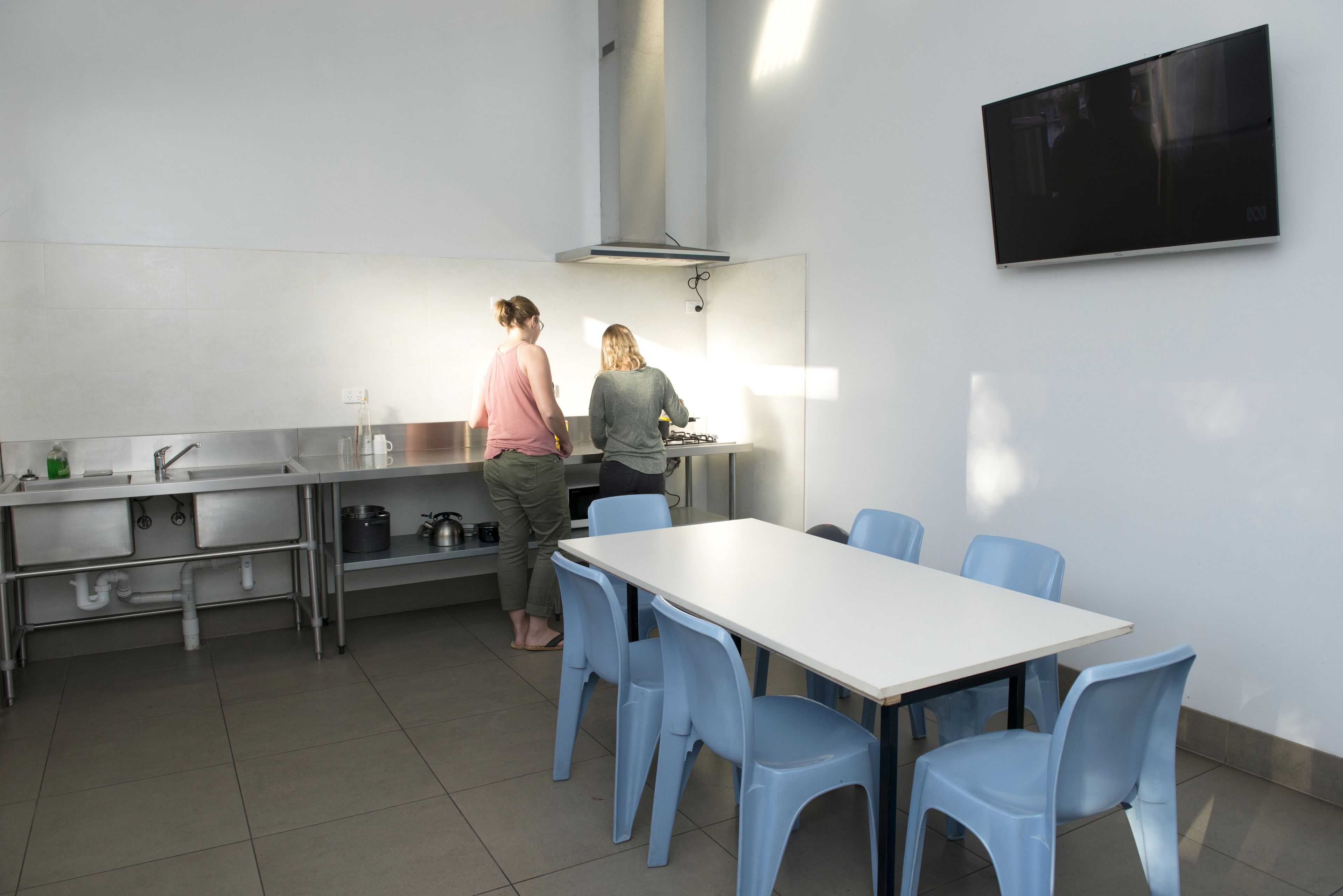 A modern kitchen with stainless steel appliances, a white tiled backsplash, and a large window providing natural light. There is a dining area with a white table and blue chairs.