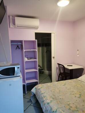 Un dormitorio pequeño y ordenado con cama, escritorio, silla, armario, microondas y aire acondicionado.