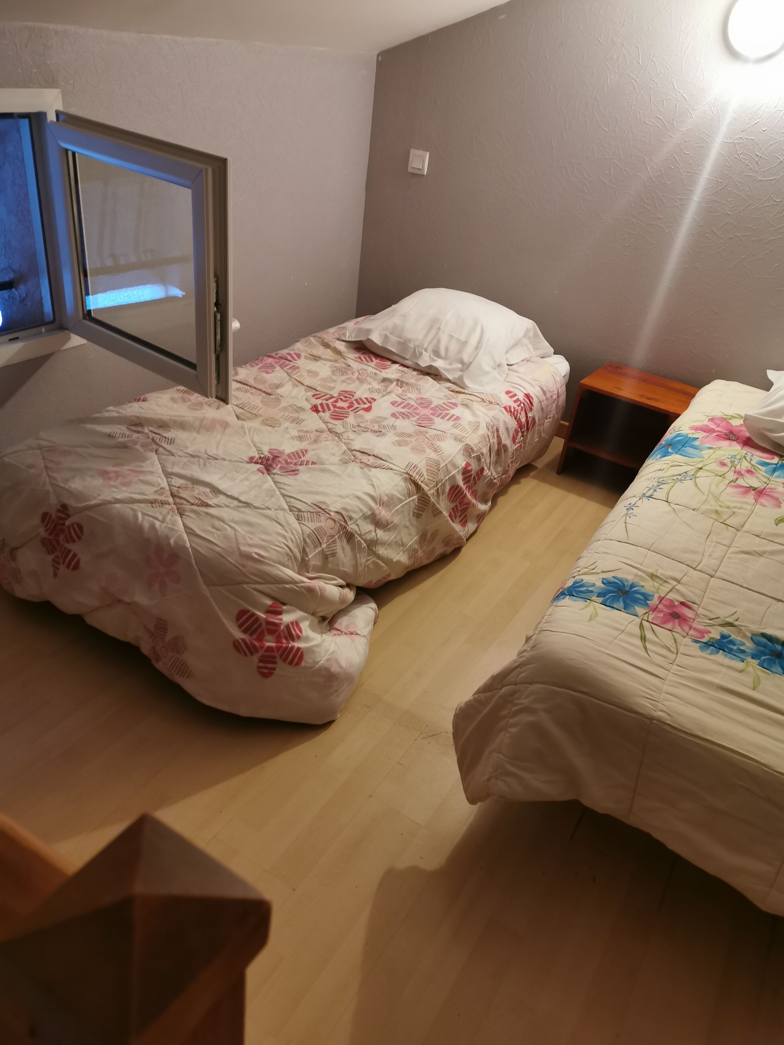 A room with two beds, one with a floral bedspread and the other with a plain one. There is a small wooden nightstand between the beds.