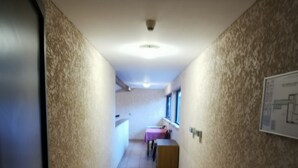 A narrow hallway with tiled flooring, a ceiling-mounted security camera, a window with blinds, and a door on the left. - Auberge de l'Europe (Saint-Quentin-Fallavier)