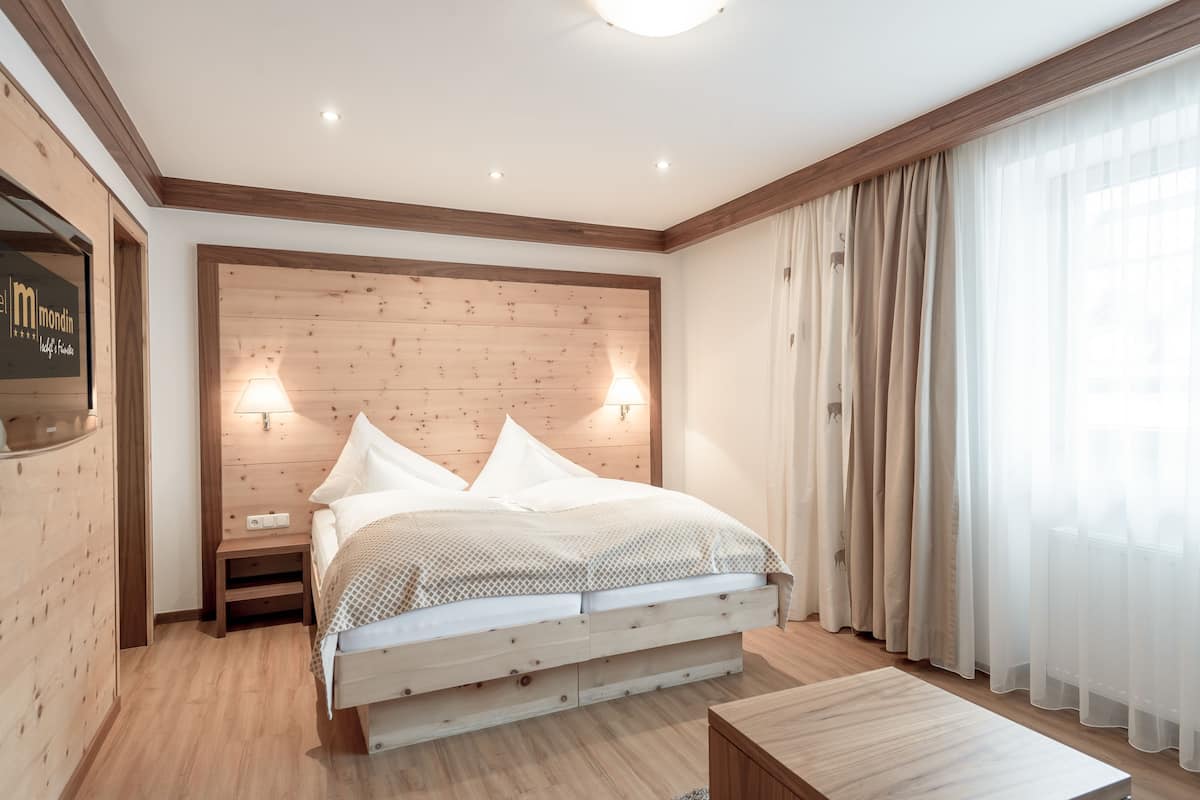 Une chambre d’hôtel moderne avec une tête de lit en bois, un lit recouvert de draps blancs, deux lampes de chevet, une table de chevet en bois et une fenêtre ornée de rideaux légers.