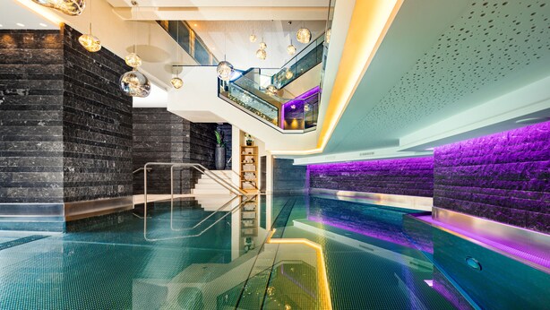 Uma piscina coberta moderna com piso de vidro, escada com corrimão de metal e parede iluminada em roxo.