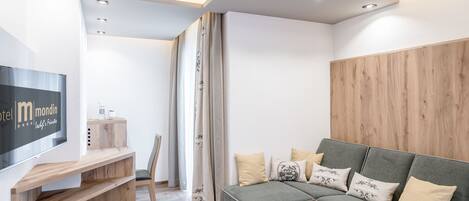 A modern hotel room with a wooden accent wall, a grey sofa with decorative pillows, a wooden coffee table, and a flat-screen TV mounted on the wall.
