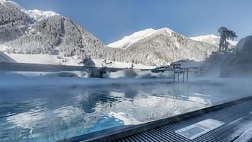 A hot spring pool with steam rising, surrounded by snow-covered mountains and trees.