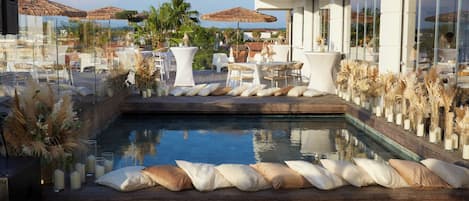 Un área de piscina en la azotea con terraza de madera, cojines blancos y baranda de vidrio.
