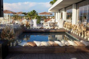 Un área de piscina en la azotea con terraza de madera, cojines blancos y baranda de vidrio.