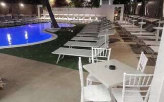 Un área junto a la piscina con sillones y mesas dispuestas alrededor de una piscina central.