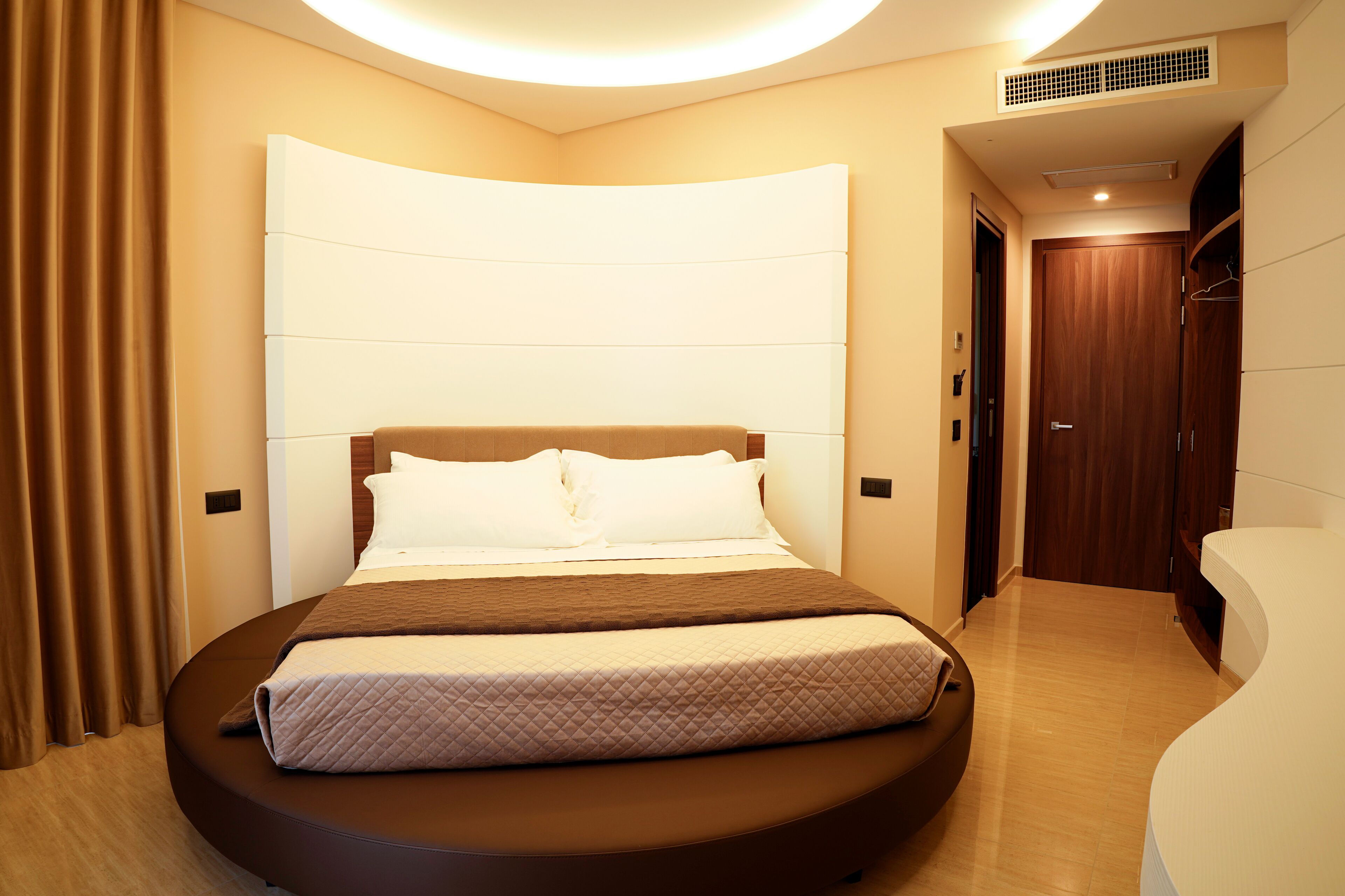 A modern hotel room with a curved bed, a large headboard, and a walk-in closet.