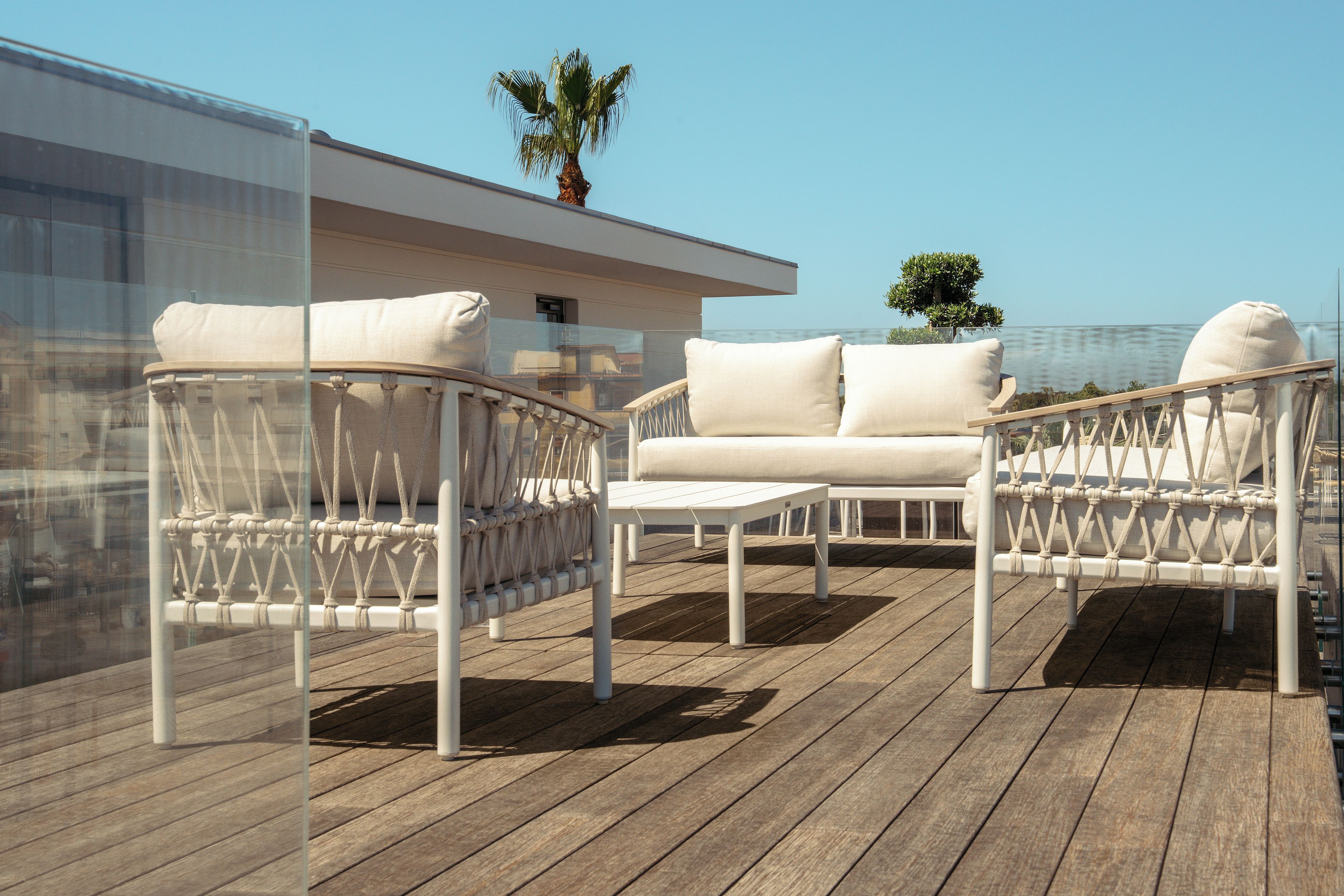 Une terrasse avec un canapé et des fauteuils blancs, un petit palmier et une vue sur la mer.