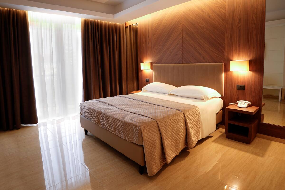 A hotel room with a large bed, wooden headboard, bedside lamps, a bedside telephone, and a large window with curtains.