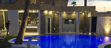 Zona de piscina con diseño moderno, que incluye una piscina de tonalidad azul, una palmera y una zona de bar con mostrador y asientos.