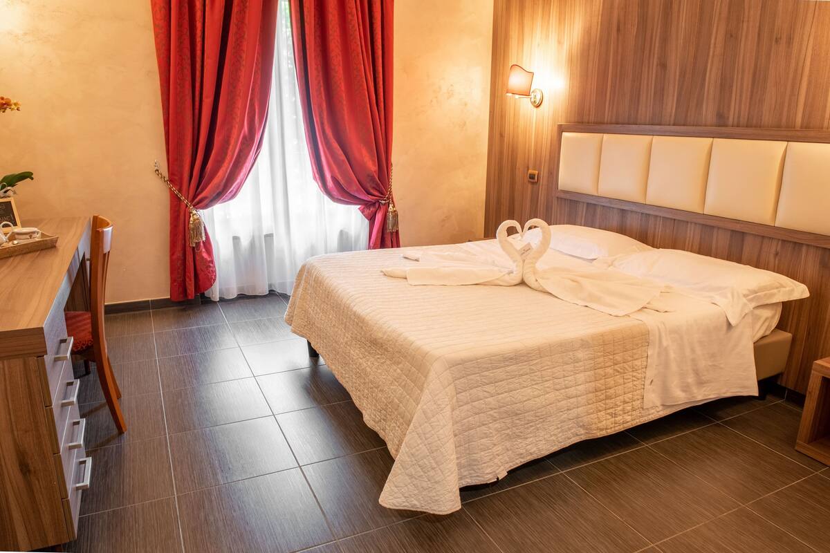 A hotel room with a bed, wooden headboard, a desk with drawers, a chair, and a window with red curtains.