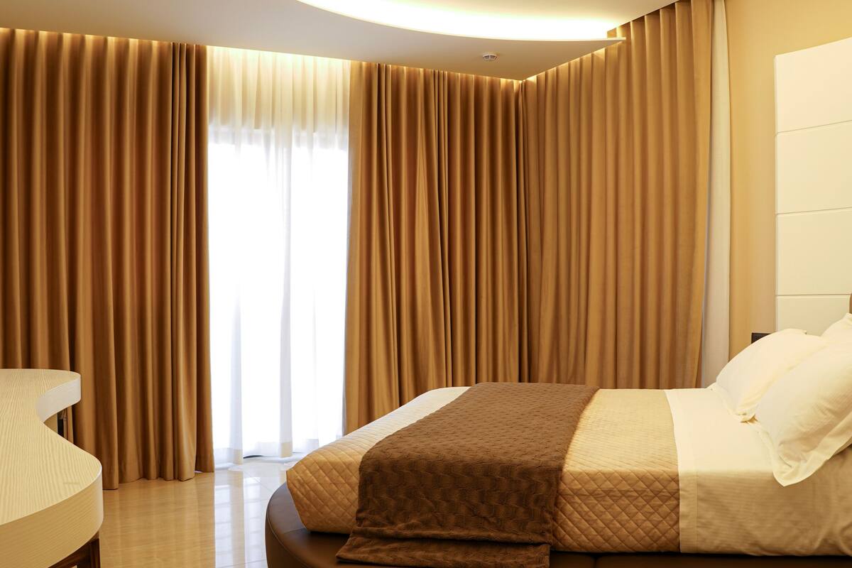 A modern hotel room with a curved bed, a round desk, and a large window with curtains.