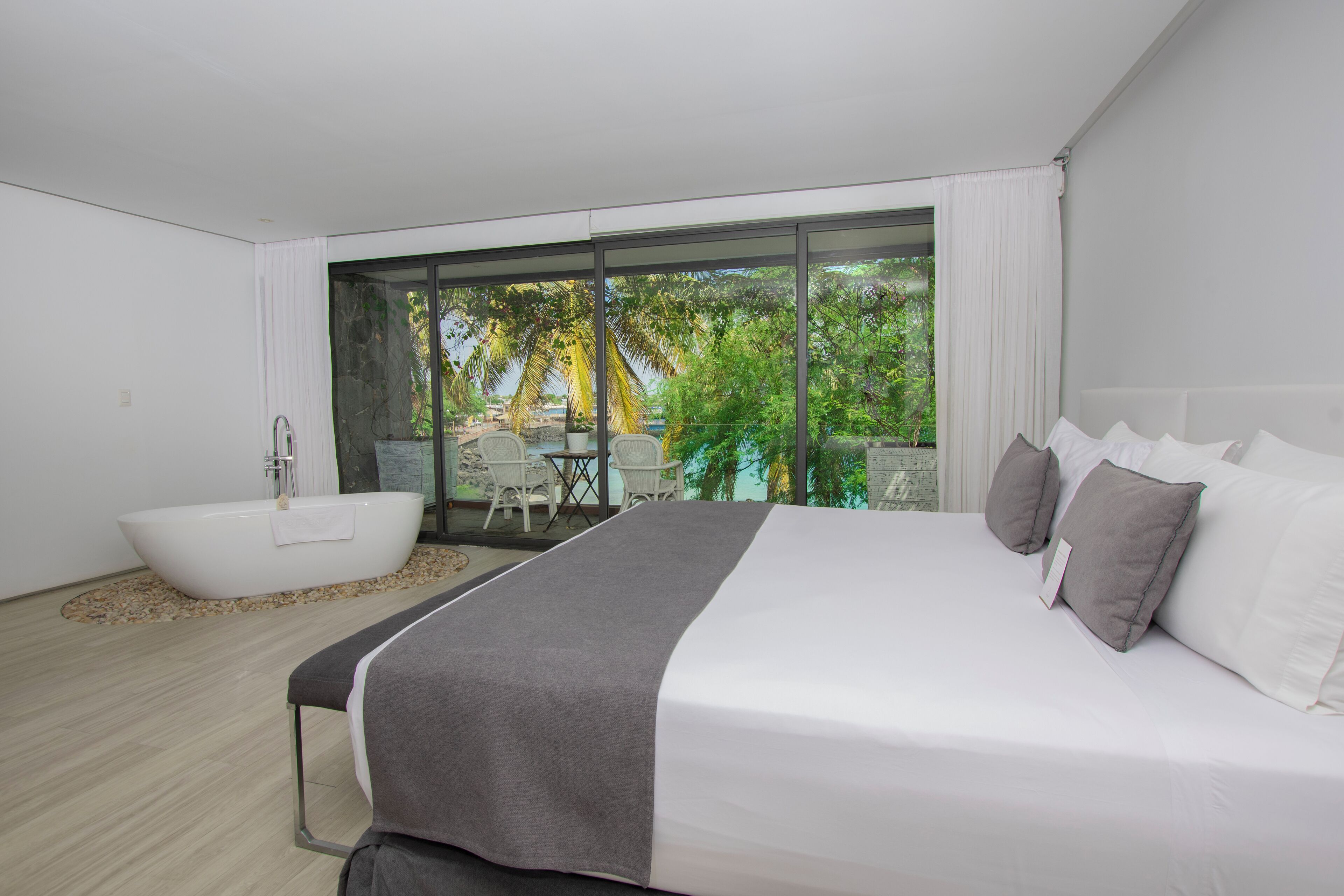 A modern bedroom with a large bed, a bathtub, and a view of a tropical garden.