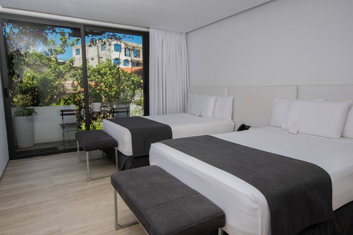 Une chambre d’hôtel moderne avec deux lits, un banc, une petite table et une vue sur les bâtiments et la verdure, grâce à de grandes fenêtres.