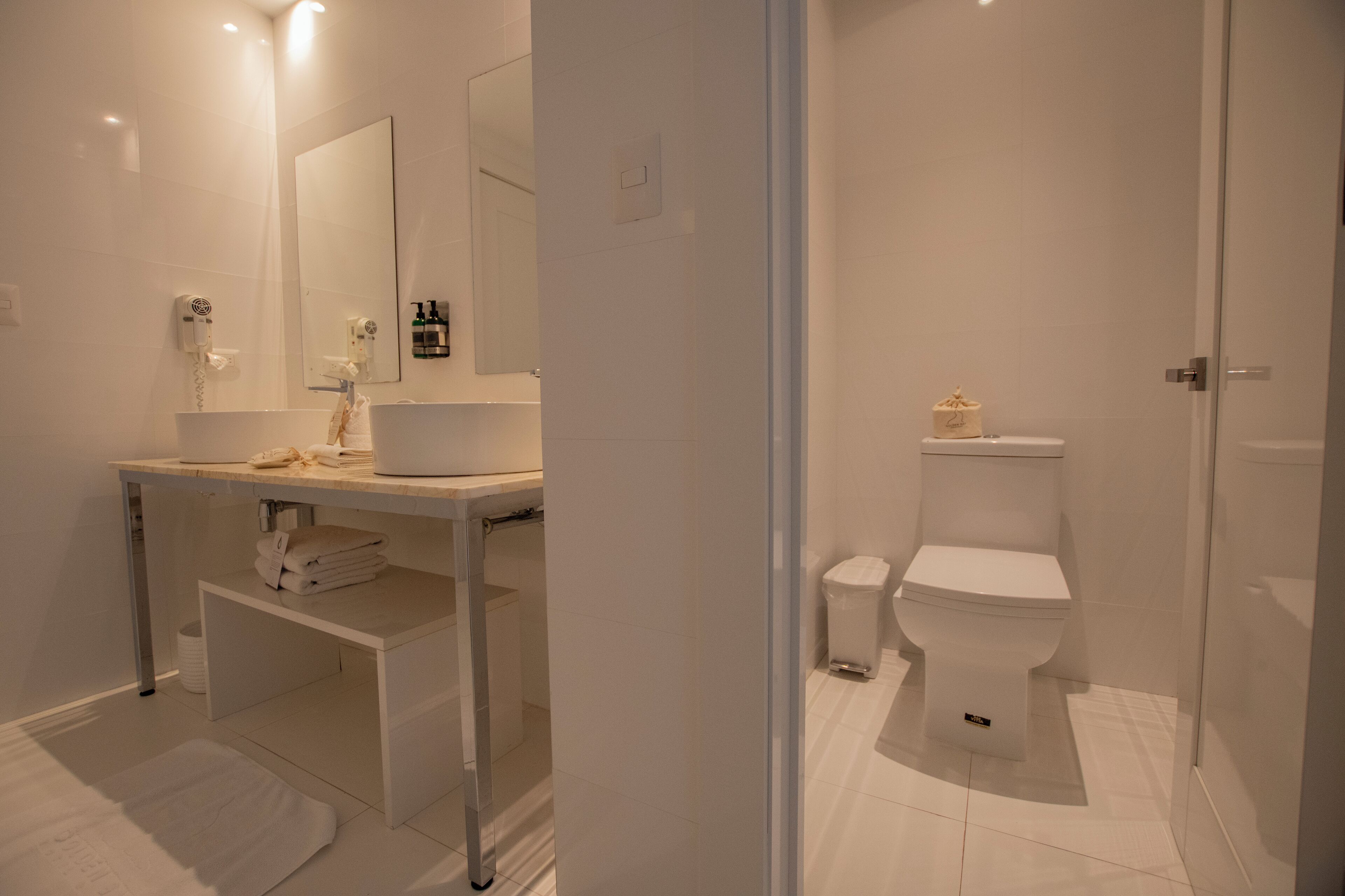 Une salle de bain moderne avec un lavabo blanc, un miroir et des toilettes. On y trouve également un porte-serviettes et une petite poubelle.