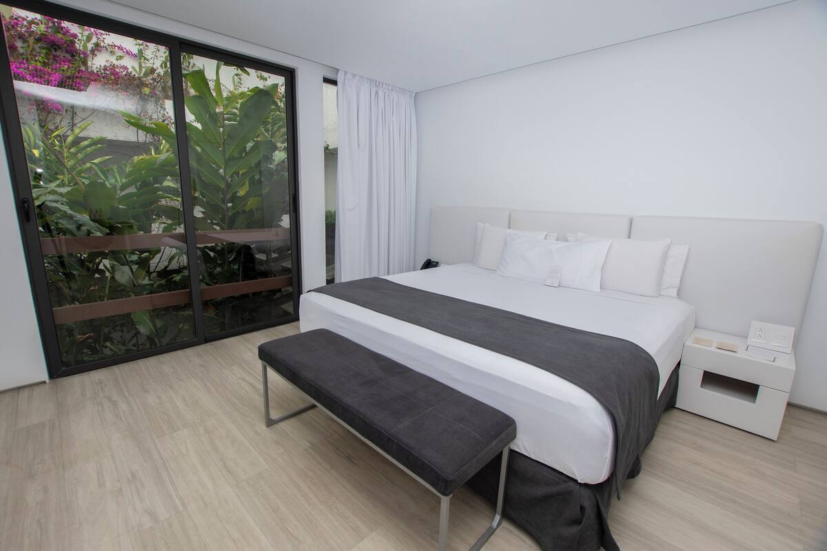 Une chambre moderne avec un grand lit, un banc, une table de chevet et une porte-fenêtre coulissante donnant sur un balcon agrémenté de plantes.