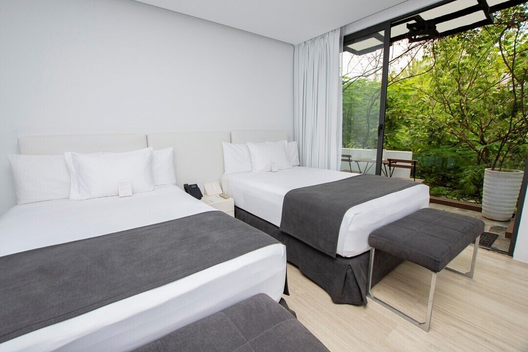 Habitación de hotel con dos camas, un banco, una mesita y vistas a zonas verdes.