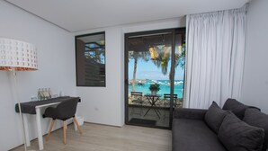 A modern living room with a dark sofa, a white desk with a chair, and a view of a beach with palm trees and boats.
