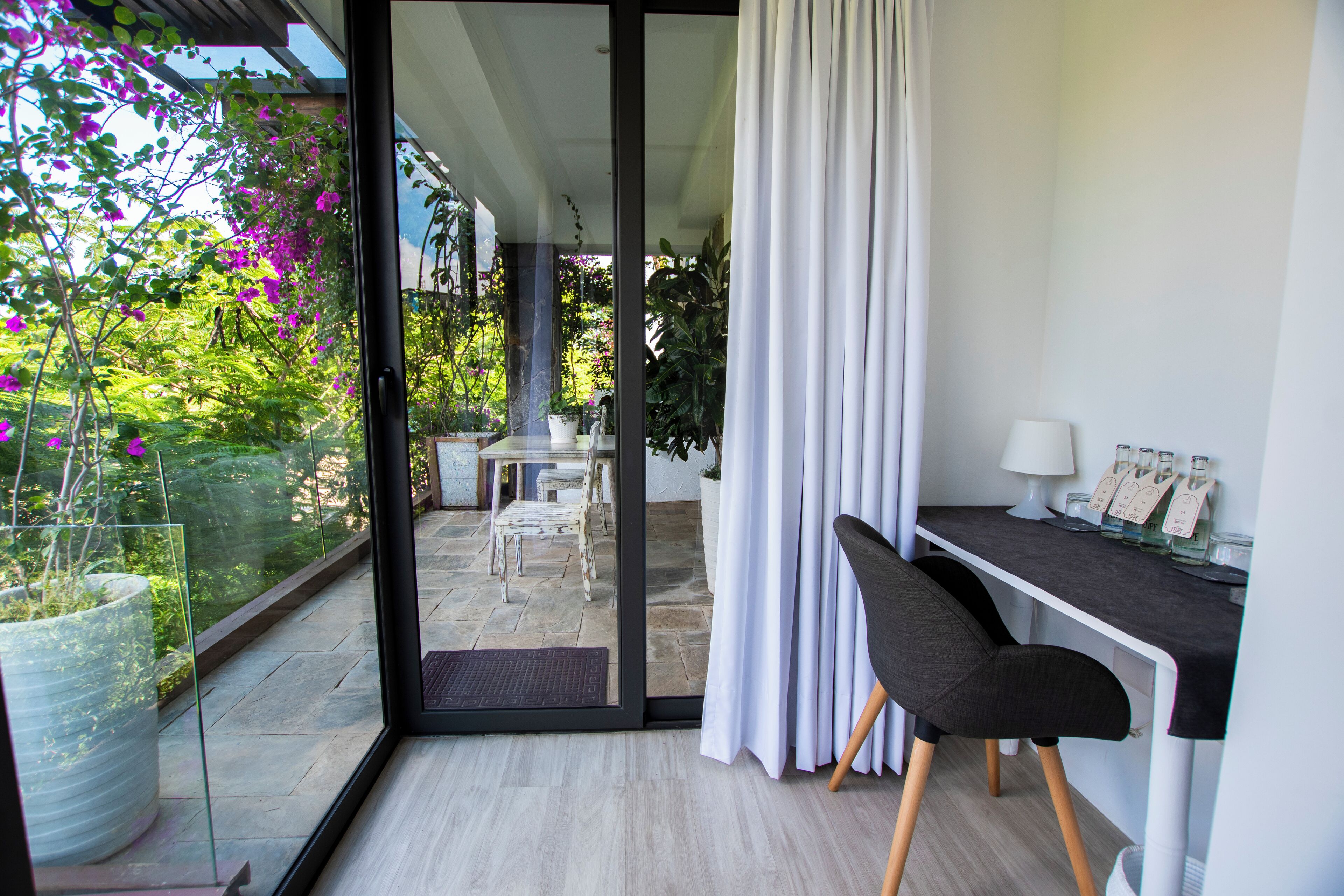 Una habitación moderna con puerta de cristal que da a un balcón, un escritorio con silla, y una pequeña mesa con sillas afuera.