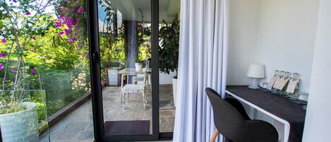 Una habitación moderna con puerta de cristal que da a un balcón, un escritorio con silla, y una pequeña mesa con sillas afuera.