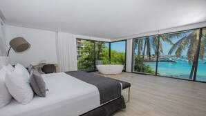 A modern bedroom with a large bed, a desk, and a view of a tropical beach and ocean.