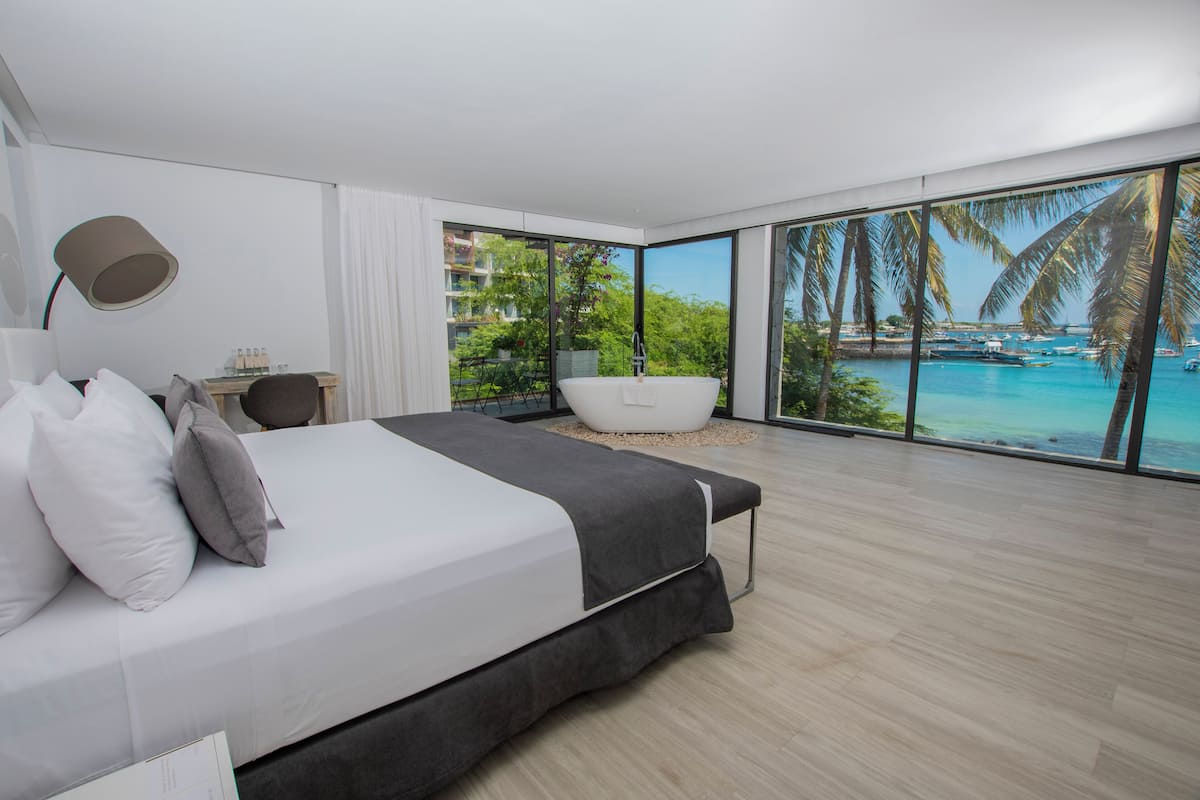 Une chambre moderne avec un grand lit, un bureau et une vue sur une plage tropicale et l’océan.