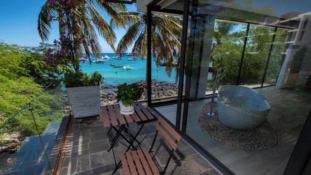 A modern outdoor seating area with a table and chairs, a large planter, and a view of the sea with boats.