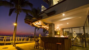 An outdoor bar area with wooden stools, a stone-tiled floor, and a view of the ocean at dusk.