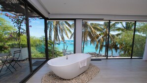 A modern bathroom with a freestanding bathtub, a wooden floor, and a view of a tropical beach and ocean through large windows.